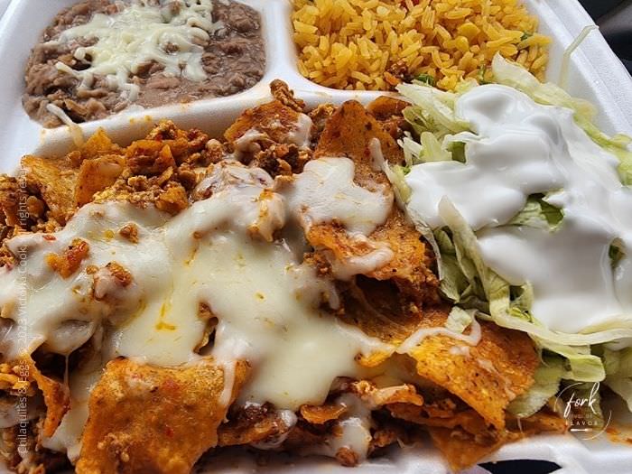 A bowl Chilaquiles and eggs made of shredded beef on top of taco chips covered with a fried egg garnished with red onion, melted shredded cheddar cheese, and cilantro sitting on a table by a cup of coffee