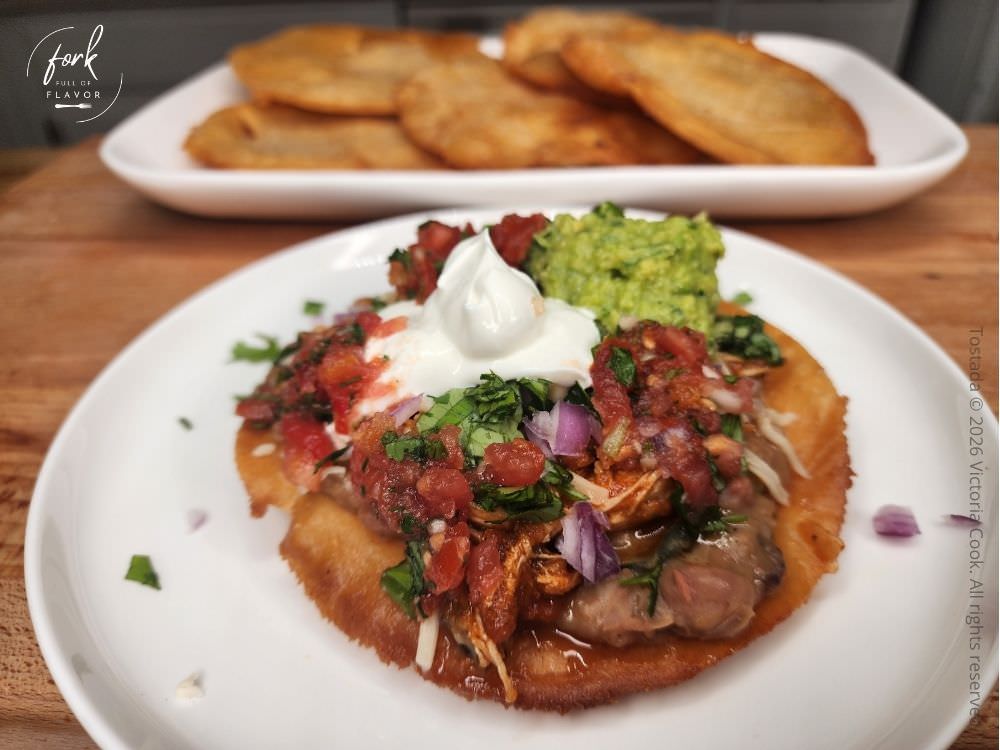 A homemade corn tostada topped with refried beans, chicken, cilantro, guacamole, and salsa.