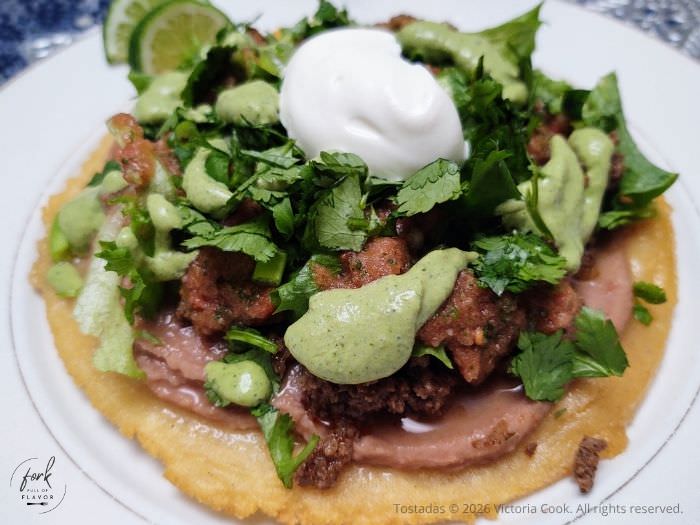 Homemade tostada shell topped with refried beans, taco meat, lettuce, sour cream and a cilantro crema.